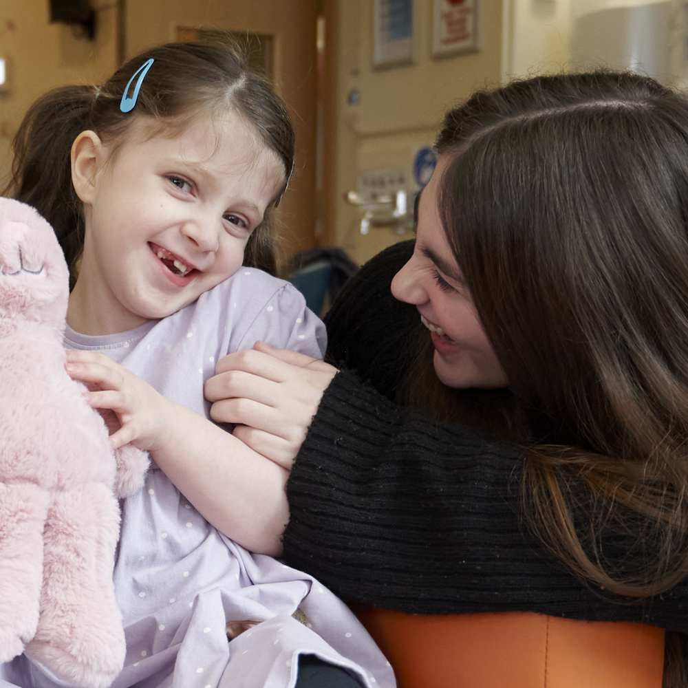 A young girl clutching a pink rabbit cuddly toy laughs with a dark haired lady in a dark jumper.