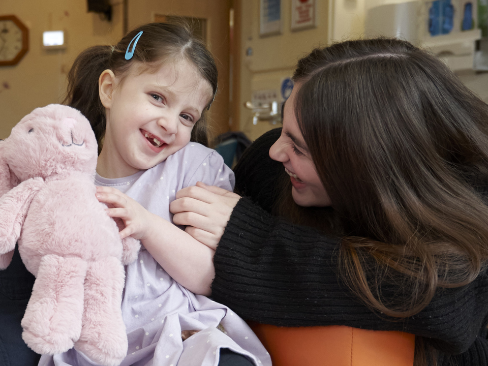 A young girl clutching a pink rabbit cuddly toy laughs with a dark haired lady in a dark jumper.